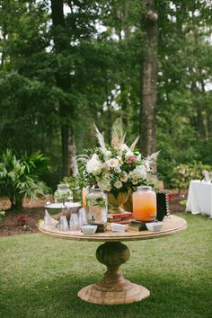 a table with candles and flowers on it in the middle of a lawn surrounded by trees