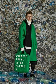 a man standing in front of a pile of plastic bottles holding a green bag that says because there is no planet b