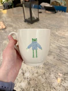 a person holding a coffee cup with a frog on it's side in front of a table