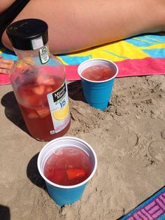 two plastic cups filled with liquid sitting on top of a beach next to a bottle