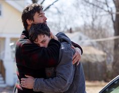 two young men hugging each other in front of a white house and tree with no leaves on it