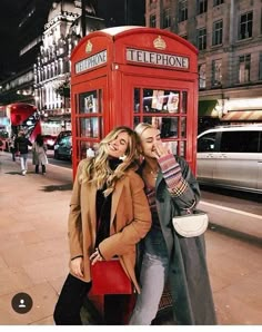 two women standing in front of a red phone booth on the side of a street