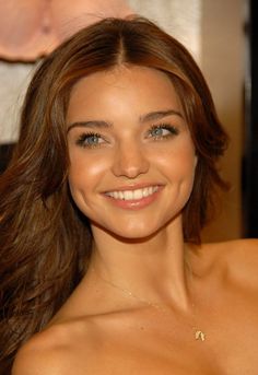 a beautiful young woman with long brown hair and blue eyes smiles at the camera while wearing a gold necklace