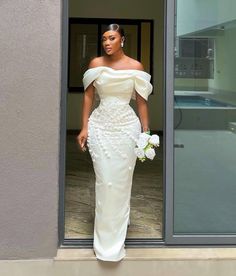 a woman standing in front of a window wearing a white dress and holding a bouquet