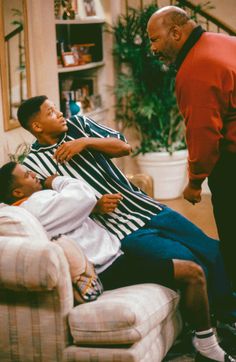 two men are talking to each other on the couch in front of a television set