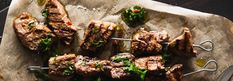 steak skewers with vegetables and sauces on a piece of parchment paper next to a knife