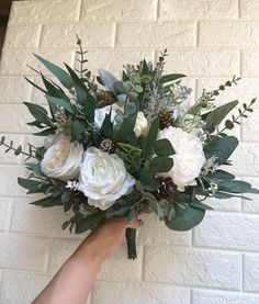 a bouquet of white flowers and greenery is held up against a brick wall by someone's hand