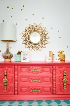 a red dresser with gold accents and a mirror above it on the wall next to a lamp