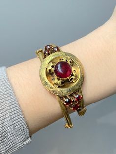 Rendered in 14K gold, this Victorian bracelet is centered with a glowing pomegranate-red garnet cabochon surrounded with scrolls of foliage leaves and motifs around. Adorned with facets of gleaming red garnets on the two sides. A work of art! Circumference of Bracelet: 6.5 inches Widest Point Measurement: 1.2 inches Measurement of Center Garnet: 0.5 inches Gold Purity: 14K Gold Markings: N/A Weight: 24.2 Grams Elegant Gold Garnet Cabochon Jewelry, Formal Yellow Gold Ruby Bangle, Elegant Ruby Bracelets For Ceremonial Occasions, Elegant Red Ceremonial Bracelets, Foliage Leaves, Victorian Bracelet, Red Pomegranate, Bottle Charms, Beautiful Perfume