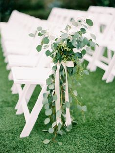 an outdoor ceremony with white folding chairs and greenery tied to the back of them