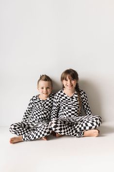 Two children sit side by side on the floor against a plain white background. They are wearing matching Bamboo Two Piece Pajamas in a Black & White Wavy Checkerboard pattern from forever french baby, made from breathable fabric. The child on the left has short hair, while the child on the right has long hair tied into two braids. Both are smiling. Wavy Checkerboard, Sack Dress, Baby Bamboo, French Baby, Play Sets, Adult Pajamas, Pajama Romper, Elastic Waistband Pants, Cozy Pajamas