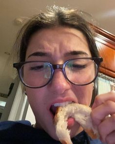 a woman wearing glasses eating a sandwich in front of her face and looking at the camera