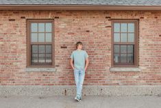 a man standing in front of two windows on a brick building with his hands in his pockets