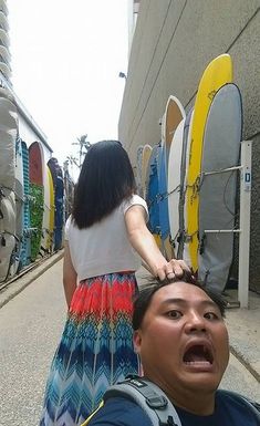 a man standing next to a woman on the street with surfboards behind him and covering his eyes