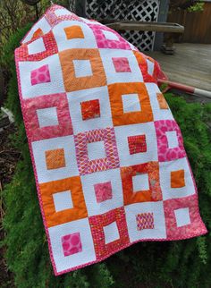 an orange and pink quilted blanket sitting on top of a wooden bench next to green grass