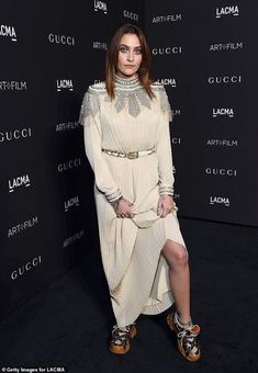 a woman standing in front of a black wall wearing sandals and a dress with high slits