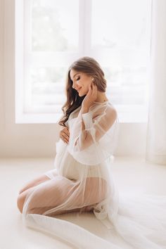 a pregnant woman sitting in front of a window wearing a white dress and holding her hands to her chest