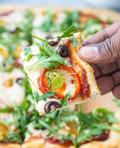 a hand holding a slice of pizza with tomatoes, olives and spinach on it