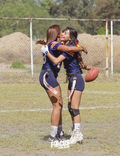 two girls are hugging each other while holding a football in front of a chain link fence