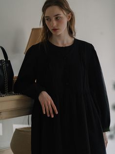 This dress exhibits a relaxed fit with a gathered waistline, creating a flared skirt that enhances its loose silhouette. It features a round neckline and a button-up front, which continues halfway down, providing a balance between structure and ease. The long sleeves are finished with elastic cuffs, adding a slight volume to the arms. The fabric looks soft and possibly lightweight, which contributes to the flowy nature of the dress.- The gathered waistline not only accentuates the dress's shape but also allows for comfortable wear.- With its simple and versatile design, this dress could be accessorized in multiple ways to suit different occasions.- The dress's button-up detail adds a classic touch, while the dark tone gives it an air of sophistication. Long Sleeve Midi Dress With Gathered Waist For Daywear, Long Sleeve Midi Dress With Gathered Waist For Work, Workwear Long Sleeve Midi Dress With Gathered Waist, Smock Dress For Workwear In Fall, Fall Smock Dress For Workwear, Fall Workwear Smock Dress, Long Sleeve Smock Dresses For Work, Black Midi Dress With Gathered Waist For Work, Black Dress With Pleated Sleeves For Daywear