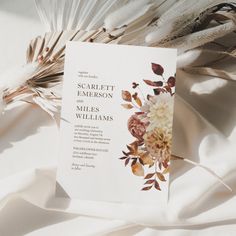 a wedding card with flowers on it laying on a white cloth covered tablecloth next to feathers