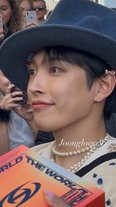 a man wearing a hat and pearls holding a book in front of his face while standing next to a group of people