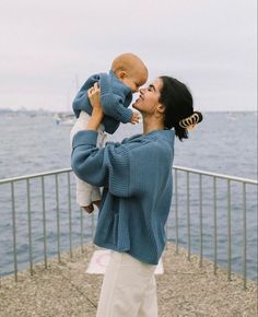 a woman holding a baby up to her face near the water with boats in the background