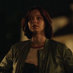 a woman standing in the dark wearing a green jacket and looking off into the distance