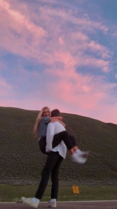 two people are hugging on the street in front of a hill and sky with pink clouds