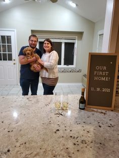 a man and woman standing in front of a counter holding a teddy bear with a sign that says our first home