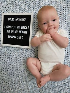 a baby is laying down next to a sign that says four months i can put my whole fist in my mouth wanna see?