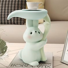 a white rabbit figurine sitting on top of a table next to a cup and saucer