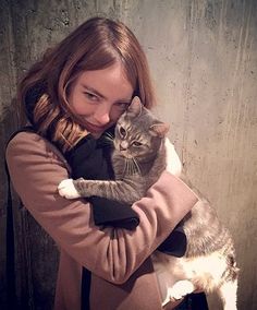 a woman holding a cat in her arms while standing next to a wall and looking at the camera