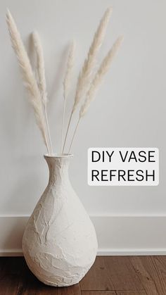 a white vase sitting on top of a wooden floor next to a wall with dry grass in it