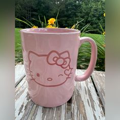 a pink hello kitty coffee mug sitting on top of a wooden table next to yellow flowers