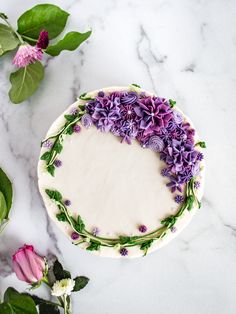 a cake decorated with purple flowers and greenery on a marble table next to green leaves