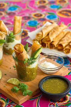 two small cups filled with food on top of a wooden cutting board next to bowls of sauce and pita chips