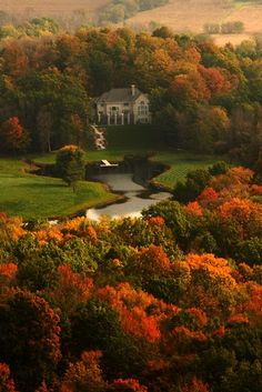 a large house surrounded by trees with fall colored leaves on the ground and hills in the background