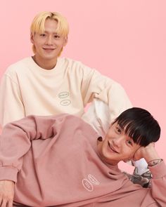 two young men sitting next to each other in front of a pink background, one wearing a sweatshirt