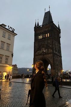 a woman walking down a cobblestone street in front of a tall clock tower