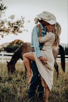 a man and woman are kissing in the grass with horses behind them, one is wearing a cowboy hat