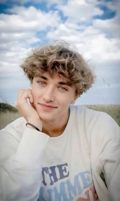 a young man with curly hair sitting in a field wearing a white shirt and looking at the camera