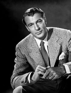 a black and white photo of a man in a suit sitting on a chair wearing a tie