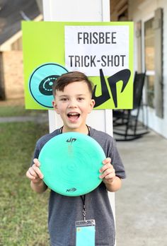 a young boy holding up a frisbee in front of a sign that says frisbee trick - shot