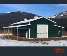 a green and white barn with snow on the ground in front of some mountain range