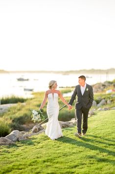 a bride and groom holding hands while walking on the grass by the water with boats in the background