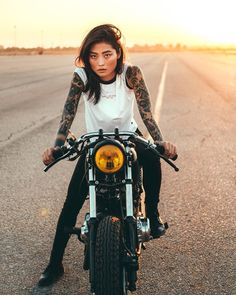 a woman sitting on top of a motorcycle in the middle of an empty road at sunset