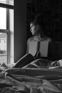 a woman sitting on top of a bed next to a window holding an open book