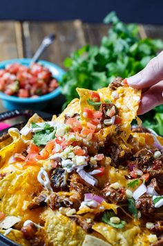 a hand holding a tortilla chip over a bowl of nachos and lettuce