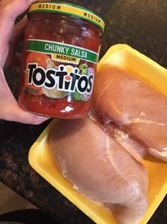 a person holding a jar of chicken salsa next to two pieces of chicken in a plastic container